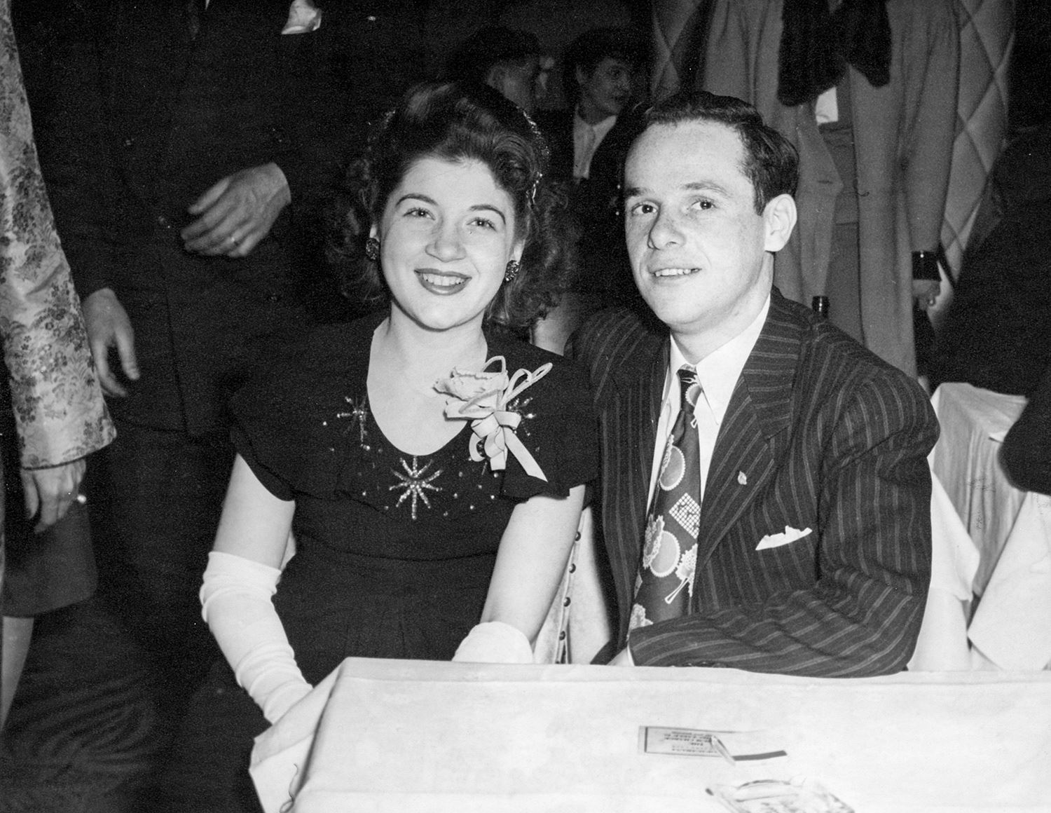 Black and white photograph of two young adults, a woman with dark hair wearing a dark dress, and a man wearing a suit and tie, smile sitting together at a table.